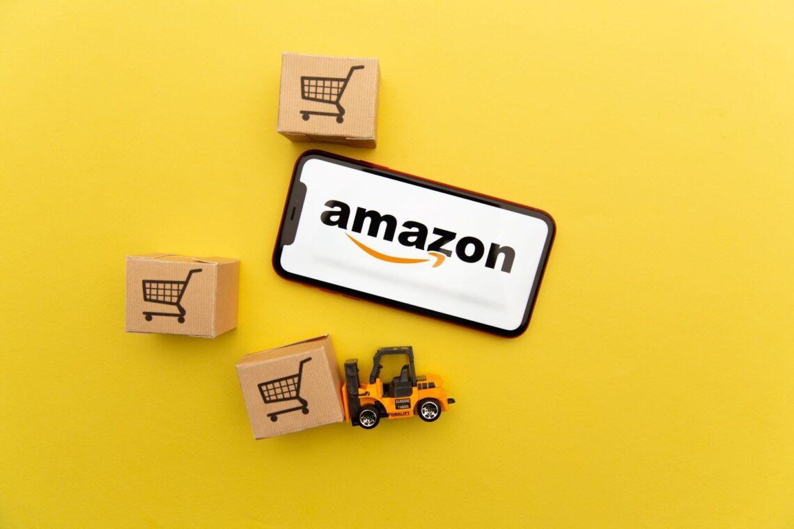 Amazon-Logo auf einem Smartphone, umgeben von Miniatur-Kartons und einem Spielzeug-Gabelstapler, auf einem gelben Hintergrund.