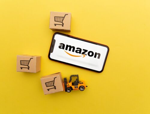 Amazon-Logo auf einem Smartphone, umgeben von Miniatur-Kartons und einem Spielzeug-Gabelstapler, auf einem gelben Hintergrund.