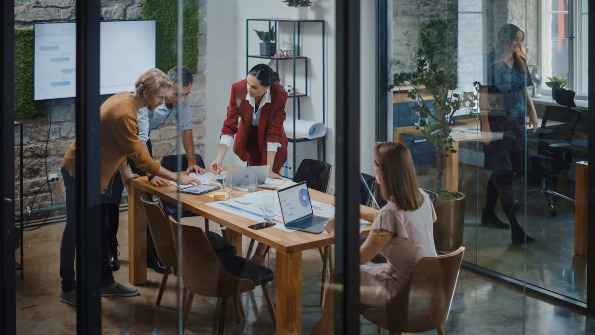 Ein Team aus vier Personen diskutiert in einem modernen Konferenzraum an einem Tisch, umgeben von Laptops und Papieren.