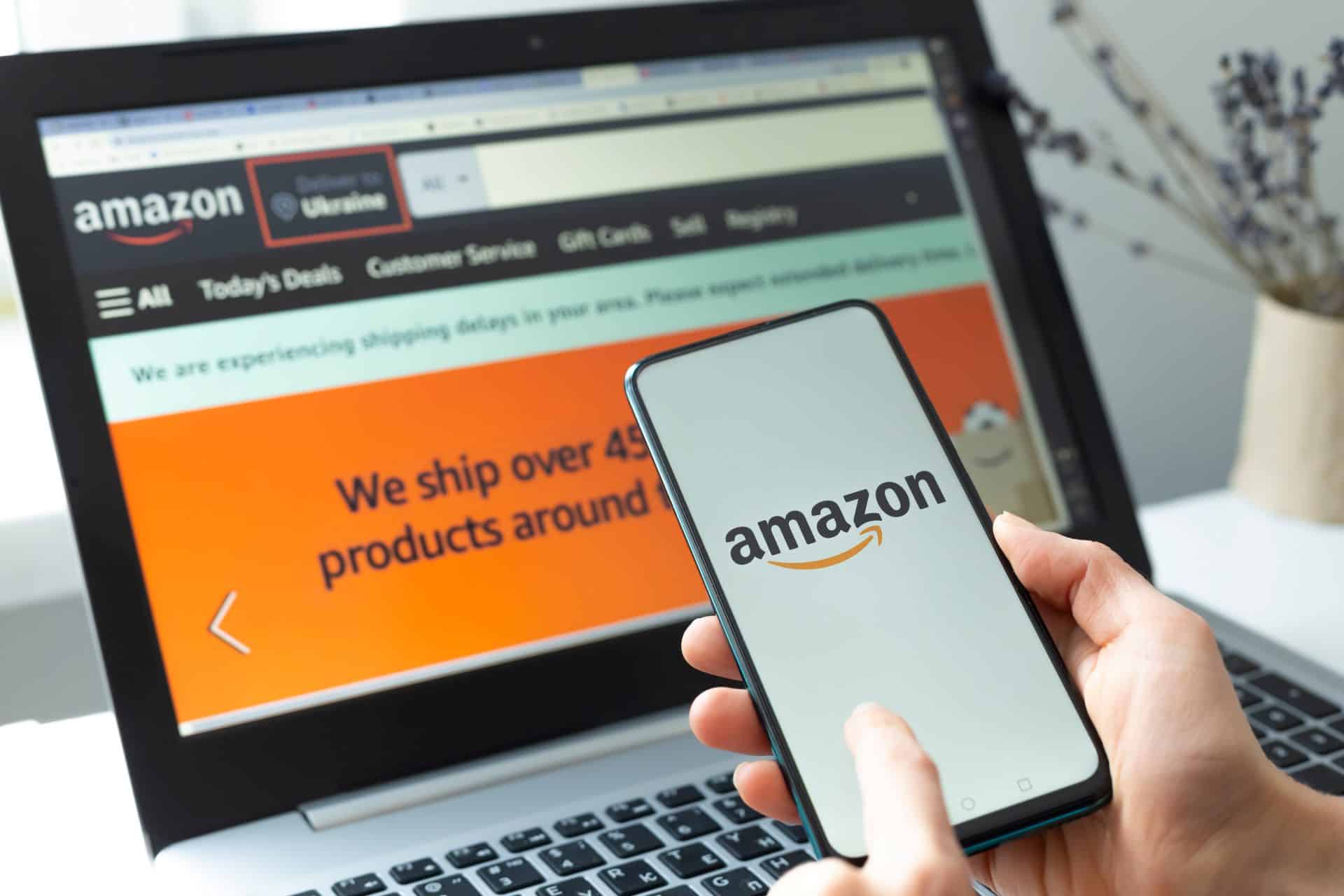 Eine Person hält ein Smartphone mit dem Amazon-Logo, im Hintergrund ein geöffneter Amazon-Shop auf einem Laptop-Bildschirm.