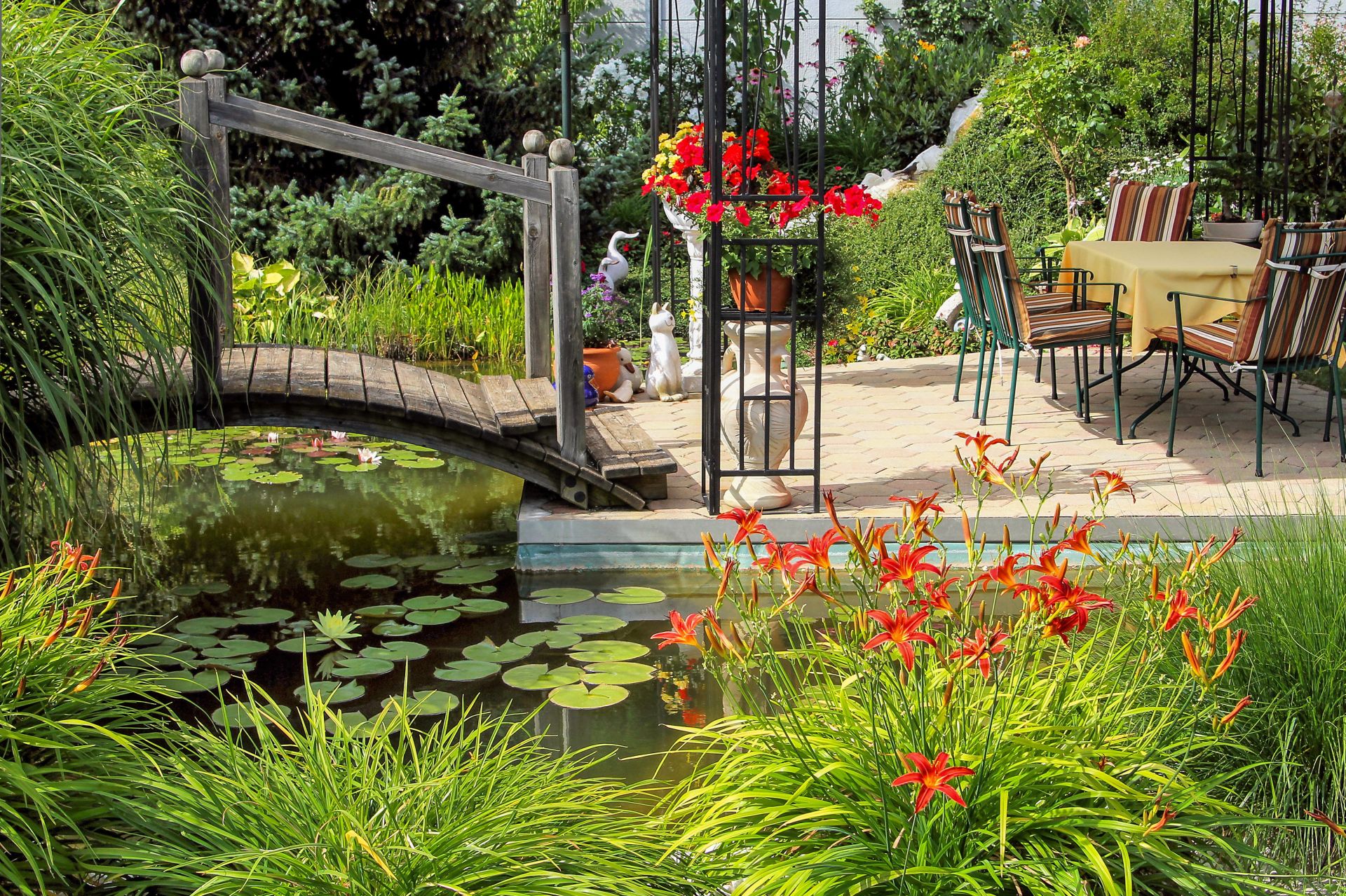 Idyllische Holzbrücke über einen Teich mit Seerosen, neben einem stilvoll eingerichteten Essbereich, umgeben von blühenden Pflanzen und Gartenpavillons.